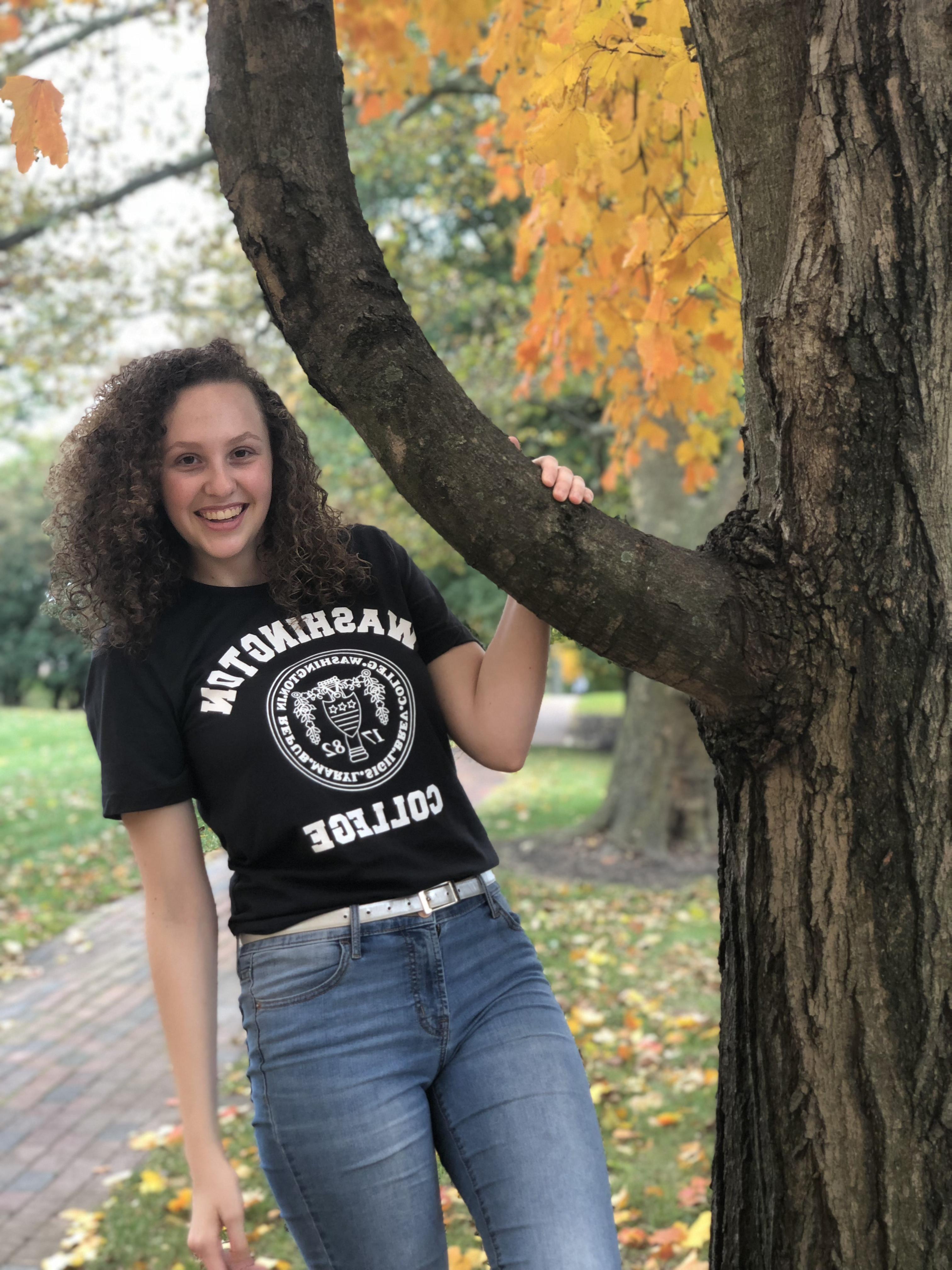 Carlee, 面带微笑，穿着印有九州娱乐官网标志的黑色t恤, 蓝色牛仔裤, 和 a white belt in front of fall foliage. 